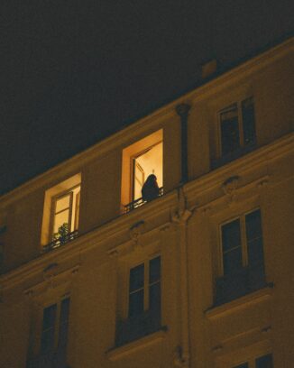 A silhouetted woman stands on a lit balcony at night, watched from the street below.
