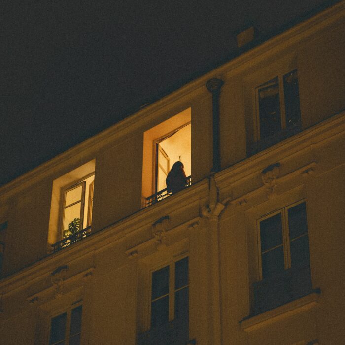 A silhouetted woman stands on a lit balcony at night, watched from the street below.