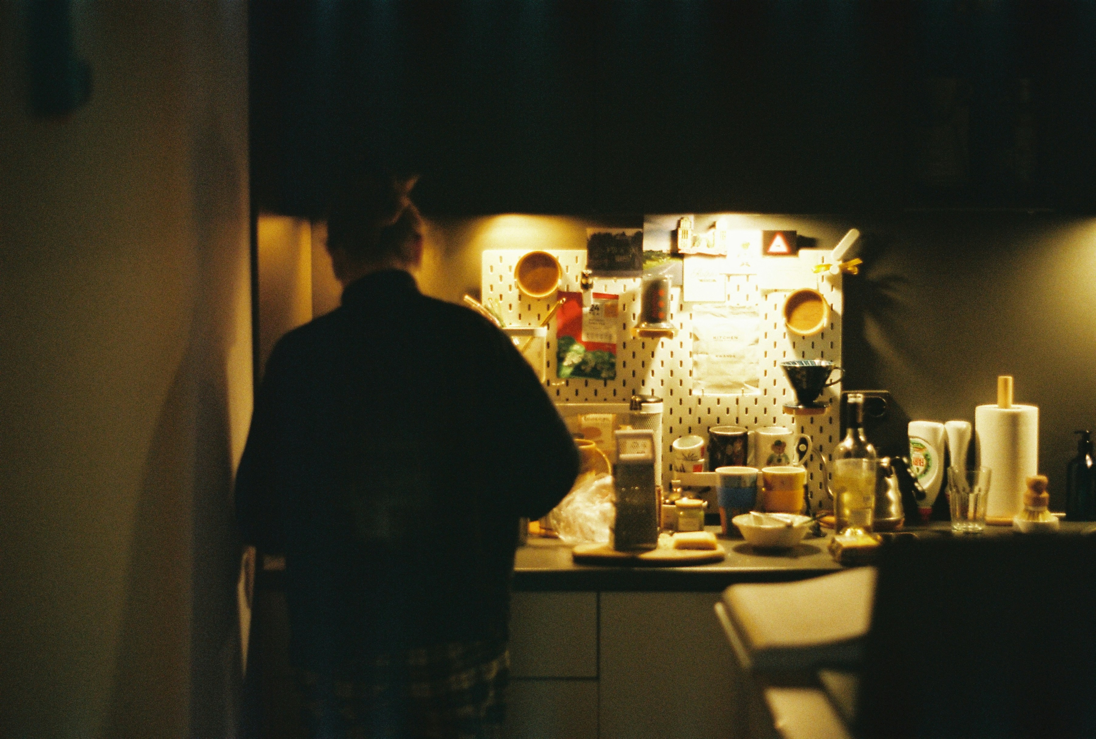 A figure standing in a dim kitchen with breakfast things scattered across the counter.