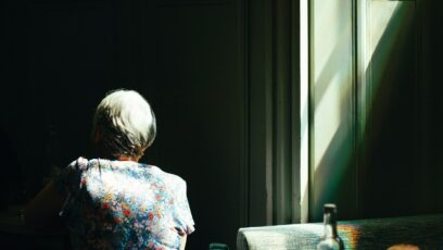 An elderly woman sitting alone at a café table beside a bright window.