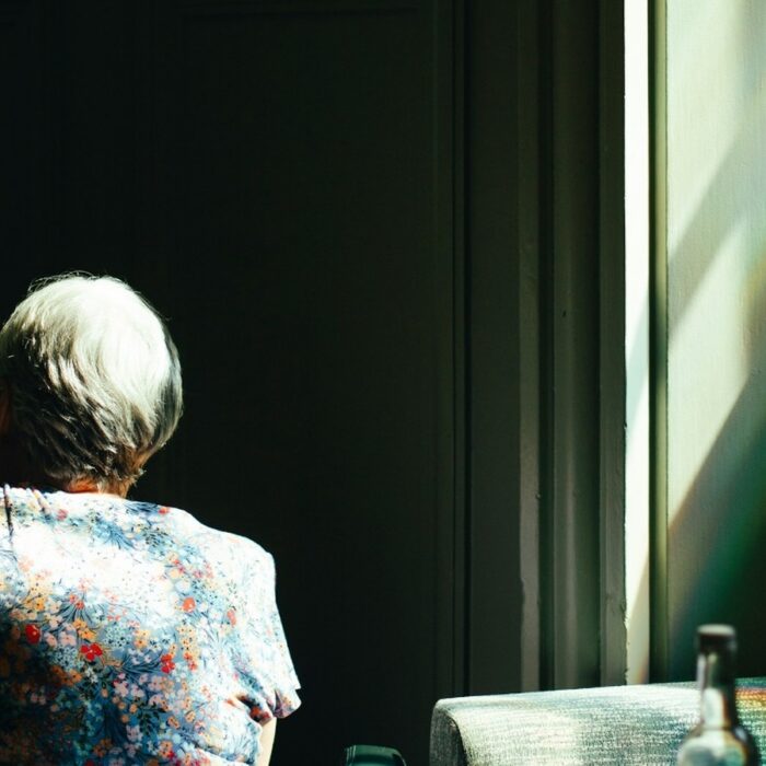 An elderly woman sitting alone at a café table beside a bright window.