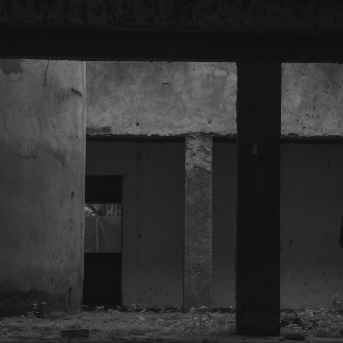 An abandoned industrial lot at night behind a chain-link fence, suggesting danger, neglect and a closed circle of violence.