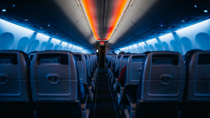 An airplane cabin aisle lit by blue side lights and a red-orange ceiling strip, suggesting an unmanned flight and approaching danger.