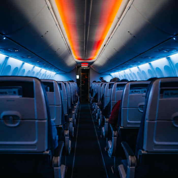 An airplane cabin aisle lit by blue side lights and a red-orange ceiling strip, suggesting an unmanned flight and approaching danger.