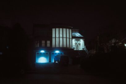 A large suburban house at night, lit by cold blue garage lights, suggesting comfort, distance and emotional unease.