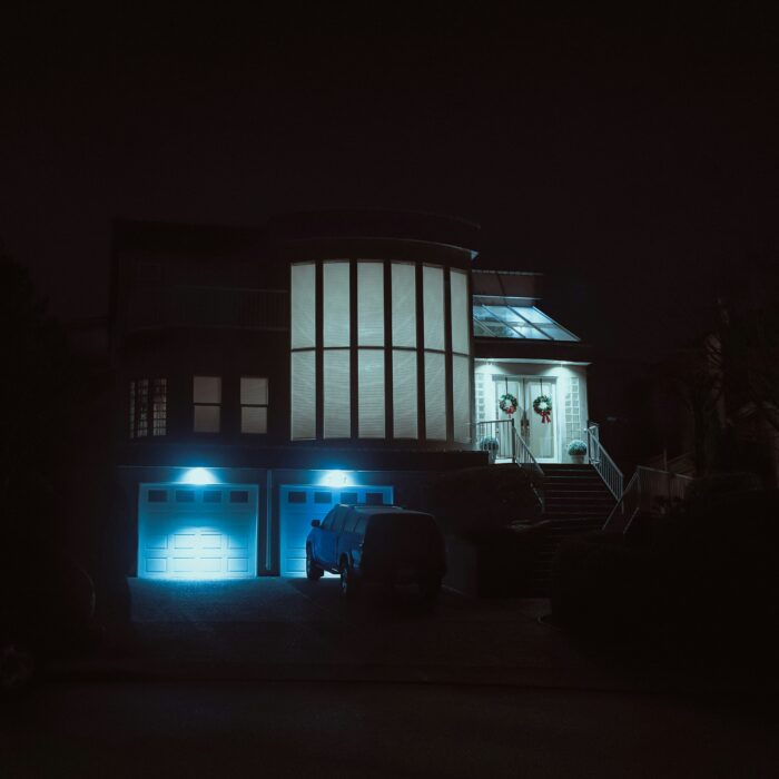 A large suburban house at night, lit by cold blue garage lights, suggesting comfort, distance and emotional unease.