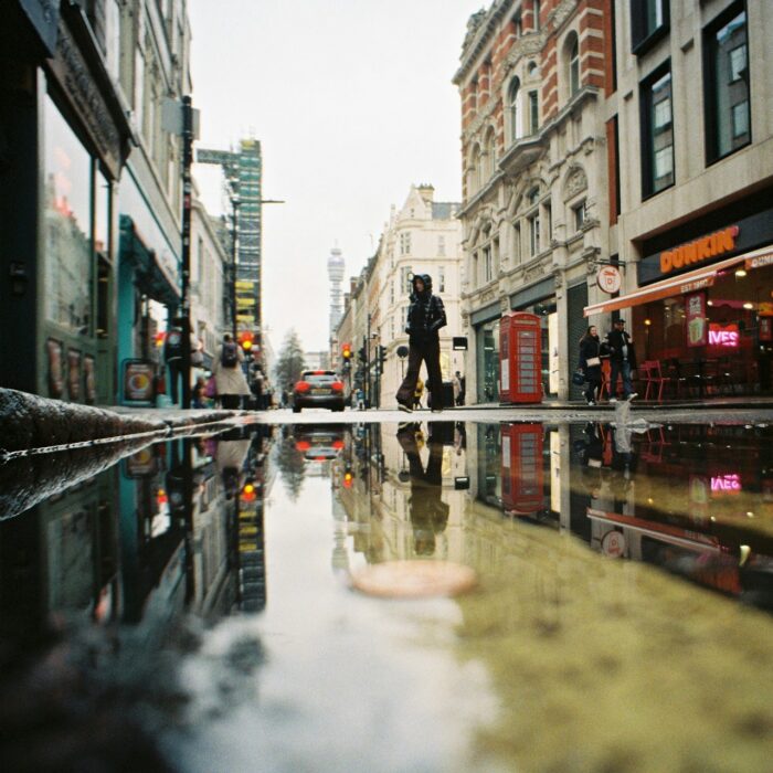 A pedestrian crossing a rain-darkened London street, reflected in a pavement puddle