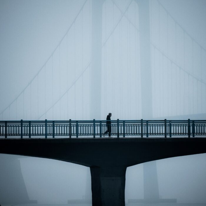 A solitary figure walks across a bridge shrouded in fog, the faint outline of suspension cables fading into the mist.