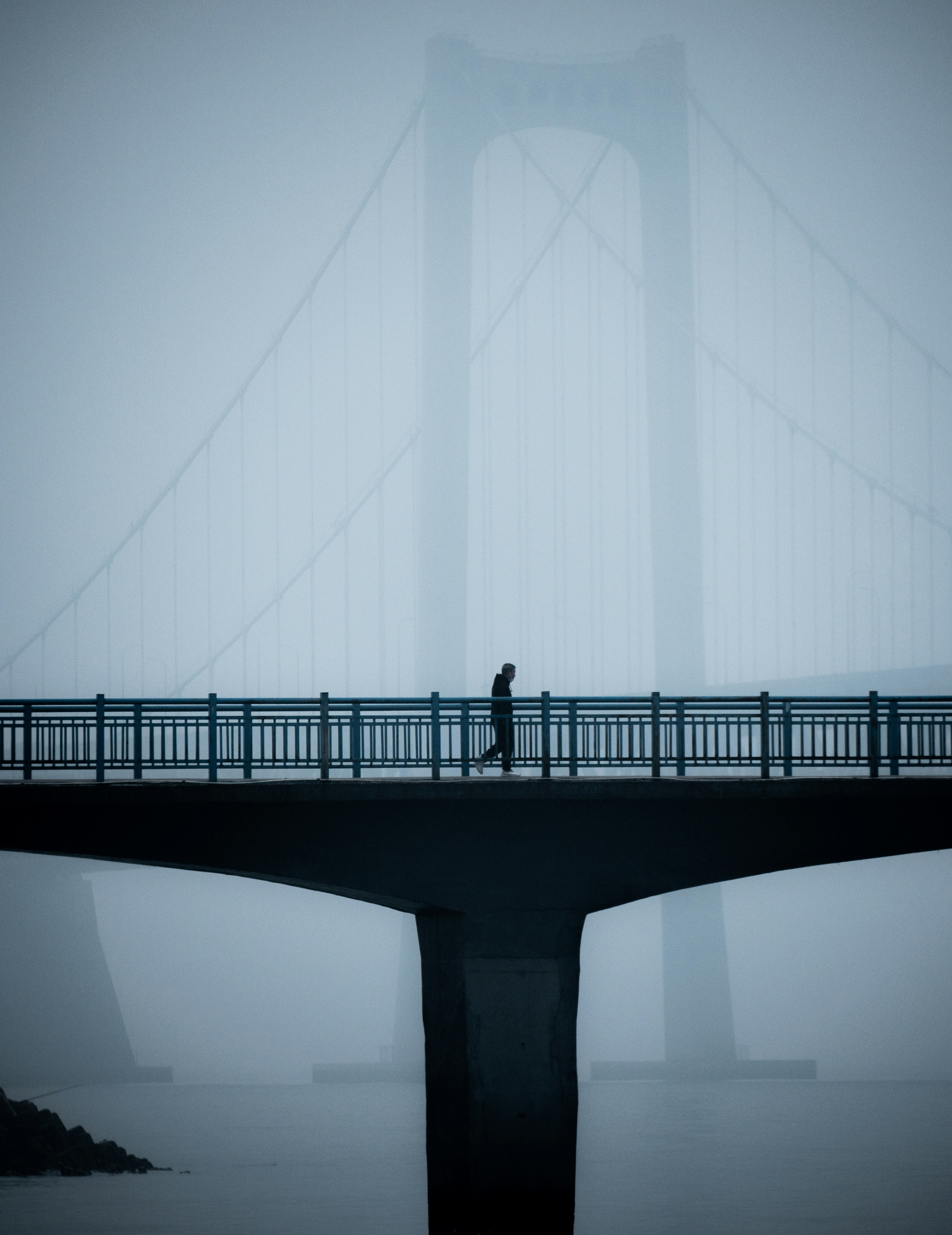 A solitary figure walks across a bridge shrouded in fog, the faint outline of suspension cables fading into the mist.