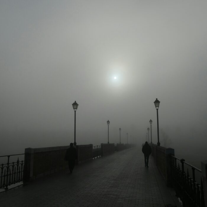 A lone figure walks across a fog-shrouded bridge under a pale sun.