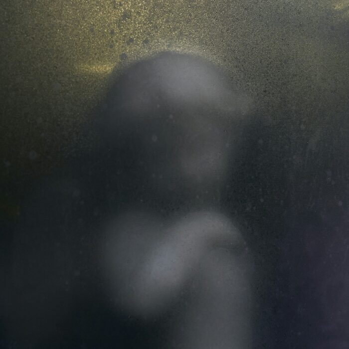 A blurred, spectral human form behind misted glass, faintly lit — evoking disintegration and artificial fragility.