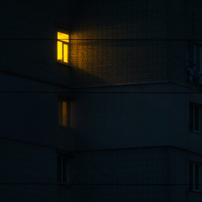 A lone lit apartment window in a dark building at night — evoking solitude, presence, and quiet unease.