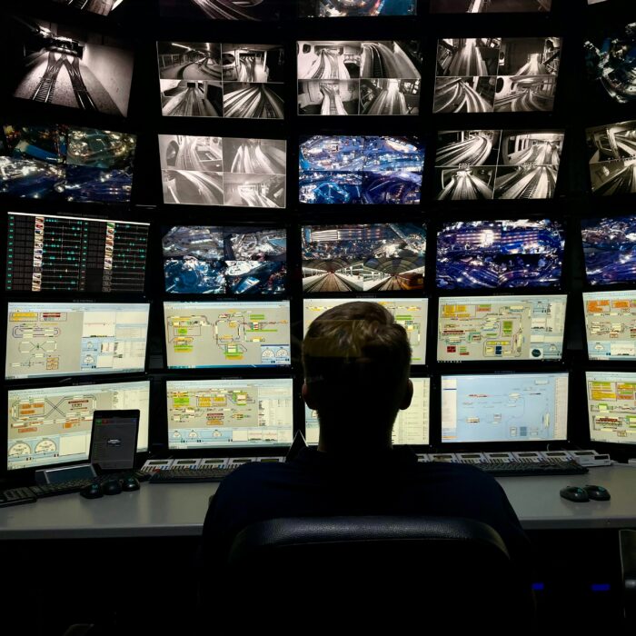 A dark surveillance control room with a seated figure facing multiple monitoring screens.