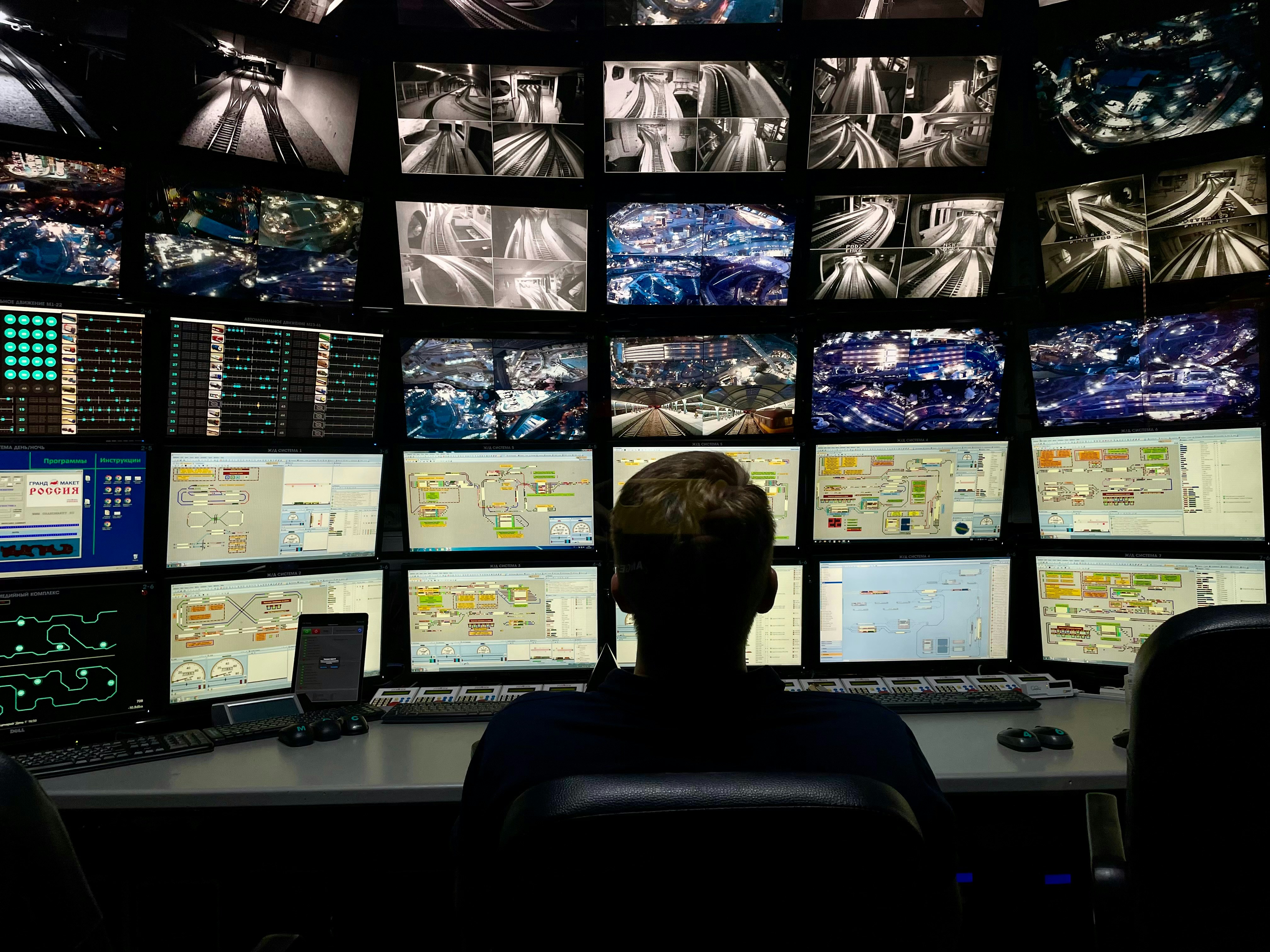 A person seated before a wall of surveillance and control-room screens.