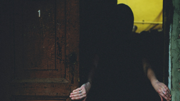 An older woman stands behind a partly open front door at dusk while a young man waits on a rain-darkened suburban path.