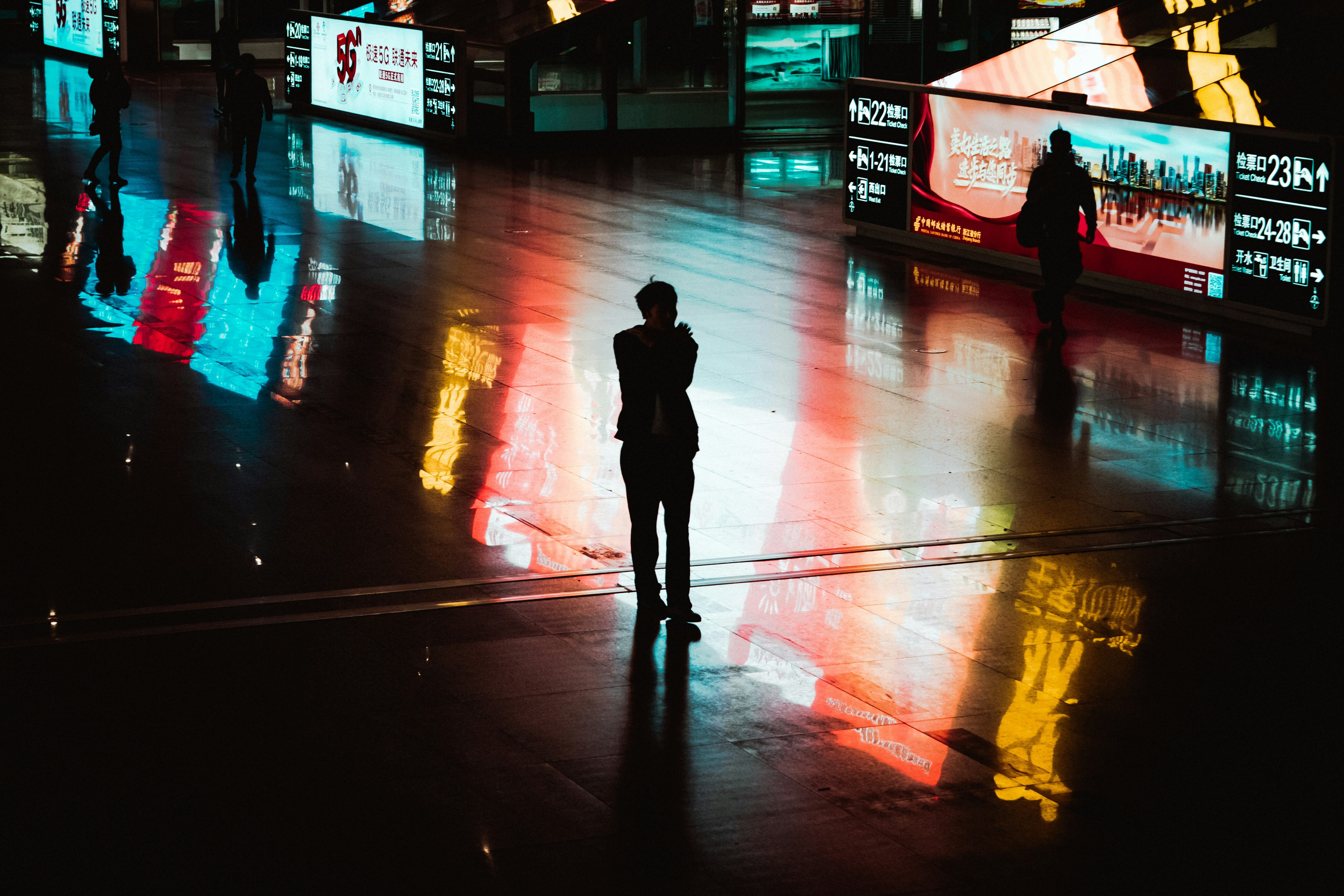A solitary figure standing inside a public space surrounded by illuminated screens and reflected information