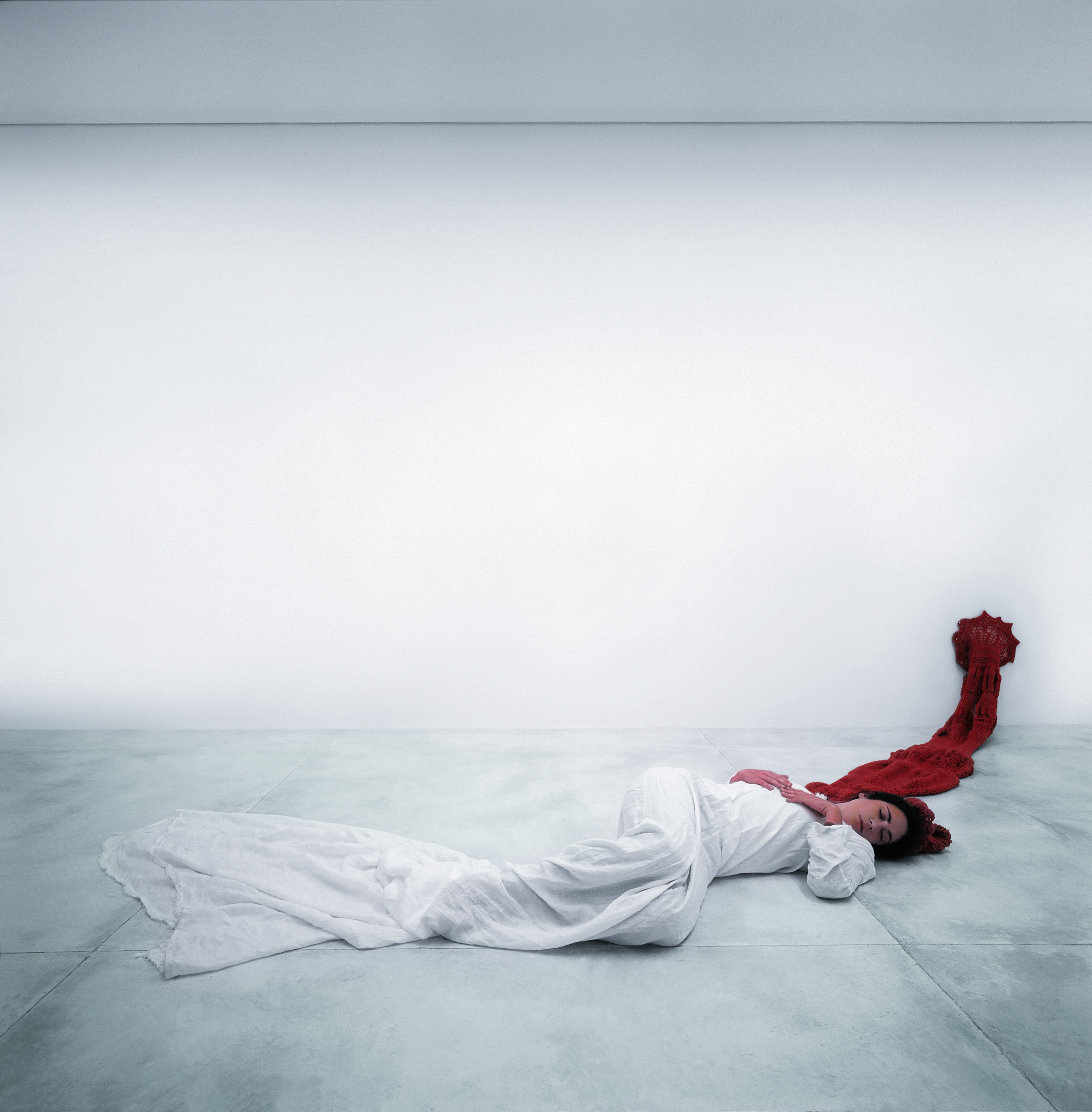 A woman lies on a pale tiled floor in a long white dress, a red crocheted form trailing from her head and climbing the gallery wall — Man=flesh/Woman=flesh — doped, by Laura Lima