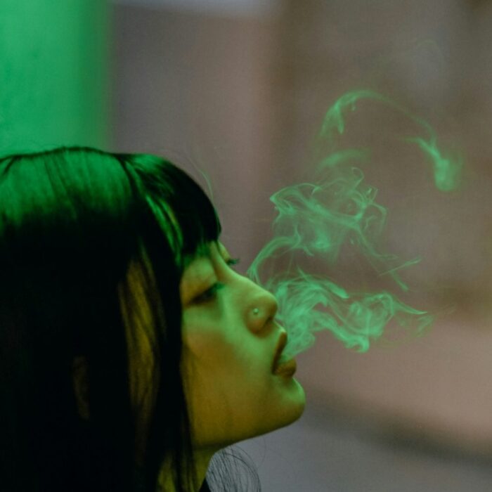 A solitary woman holding an unlit cigarette outside a rain-darkened institutional entrance at dusk.