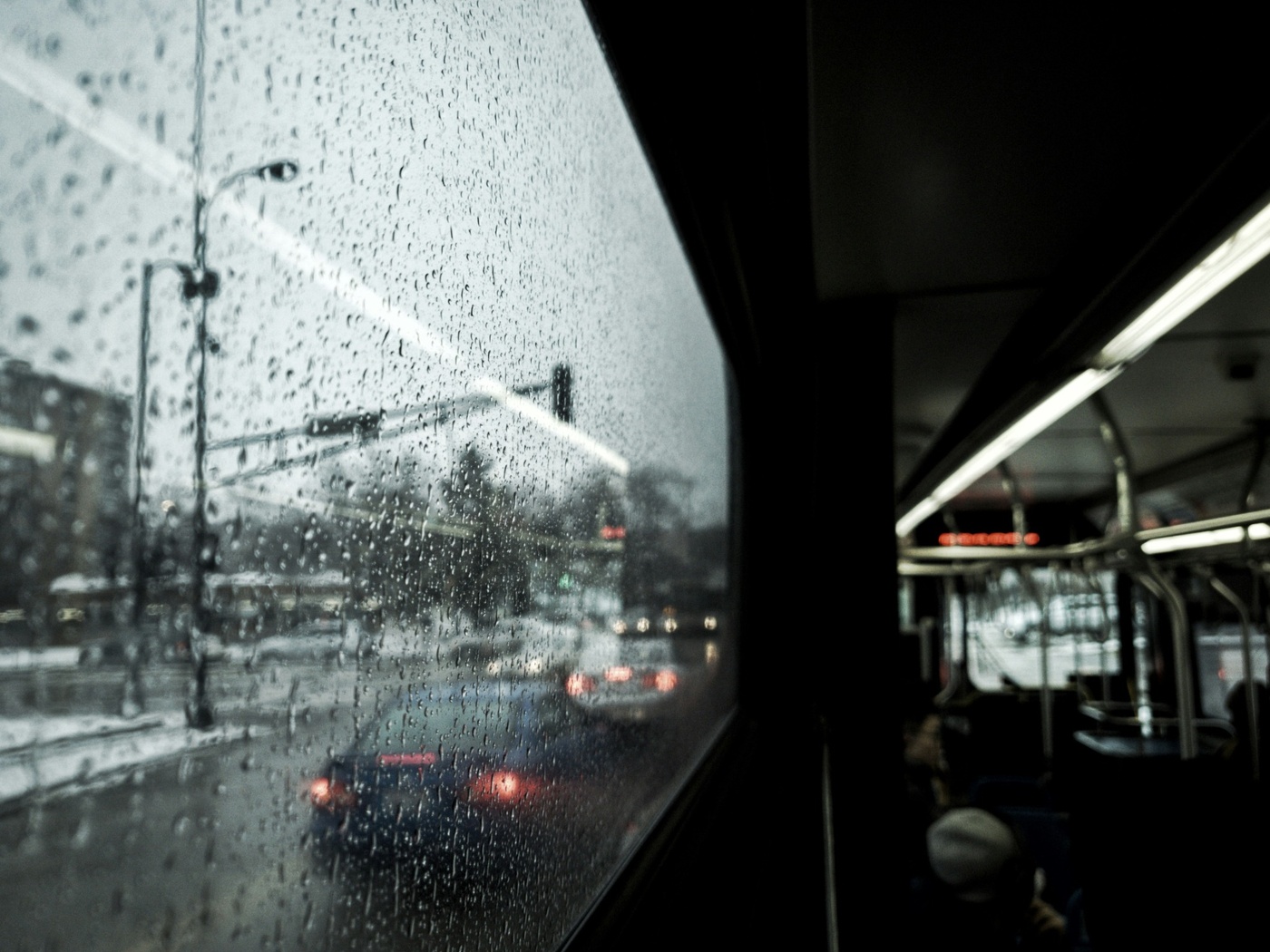 Rain streaks across a bus window, with a blurred city street outside and the dark interior of the bus reflected beside it.