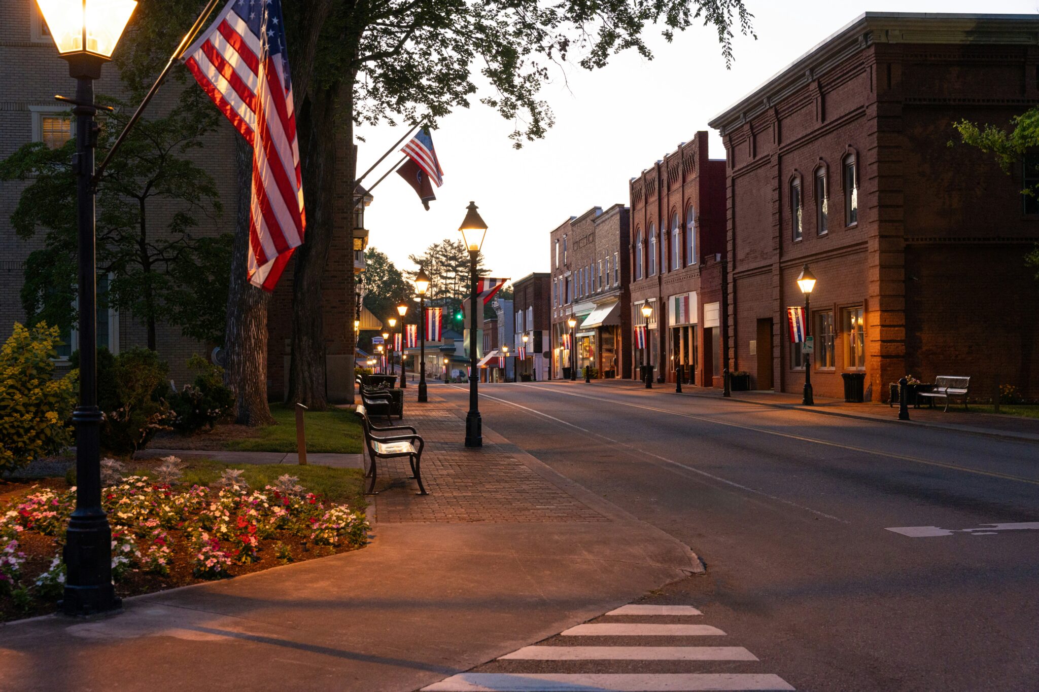 A small-town main street at dusk, lamps lit, flags hanging still