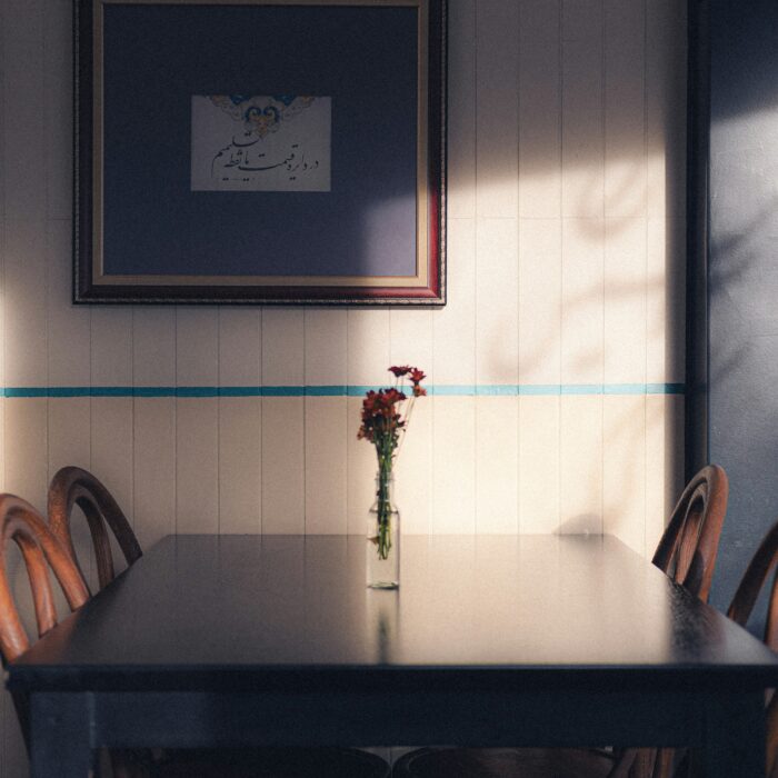 An empty dining table with wooden chairs and a small vase of flowers, lit by afternoon shadows.