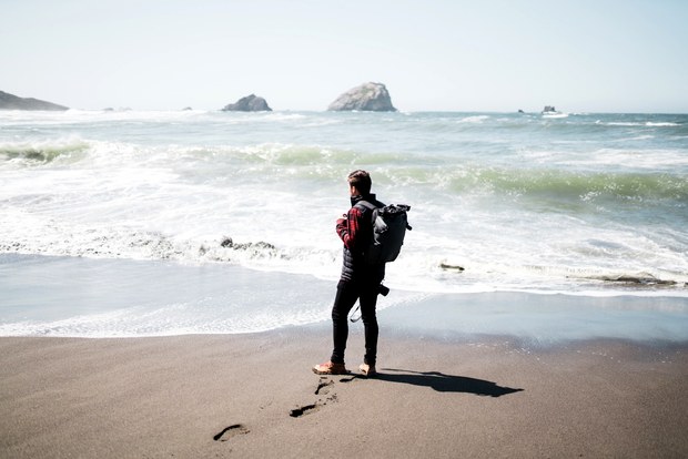 A traveller wearing a Stormtech backpack stands on a beach facing the sea.