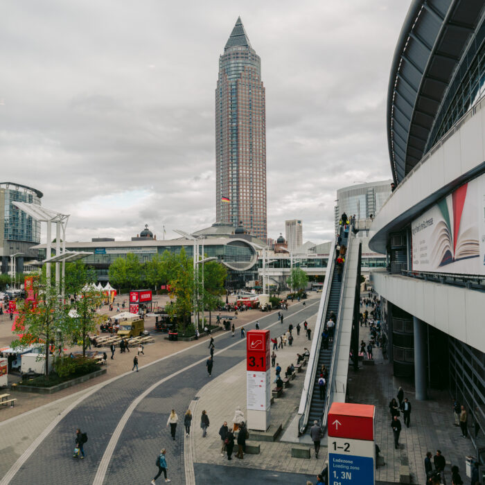 Visitors cross the Agora plaza at Frankfurter Buchmesse 2026, with the Messeturm tower and an exhibition hall behind