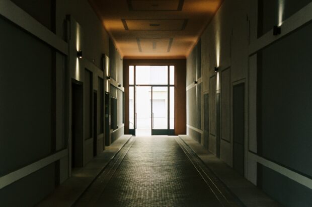A dim entrance corridor in a shared building, closed doors on either side, leading towards a bright glass door standing open.
