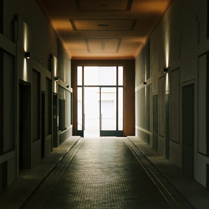 A dim entrance corridor in a shared building, closed doors on either side, leading towards a bright glass door standing open.